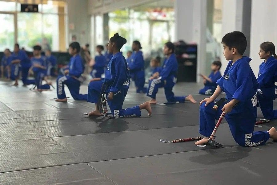 Youth class kneeling with training sticks during a disciplined martial arts weapons lesson.