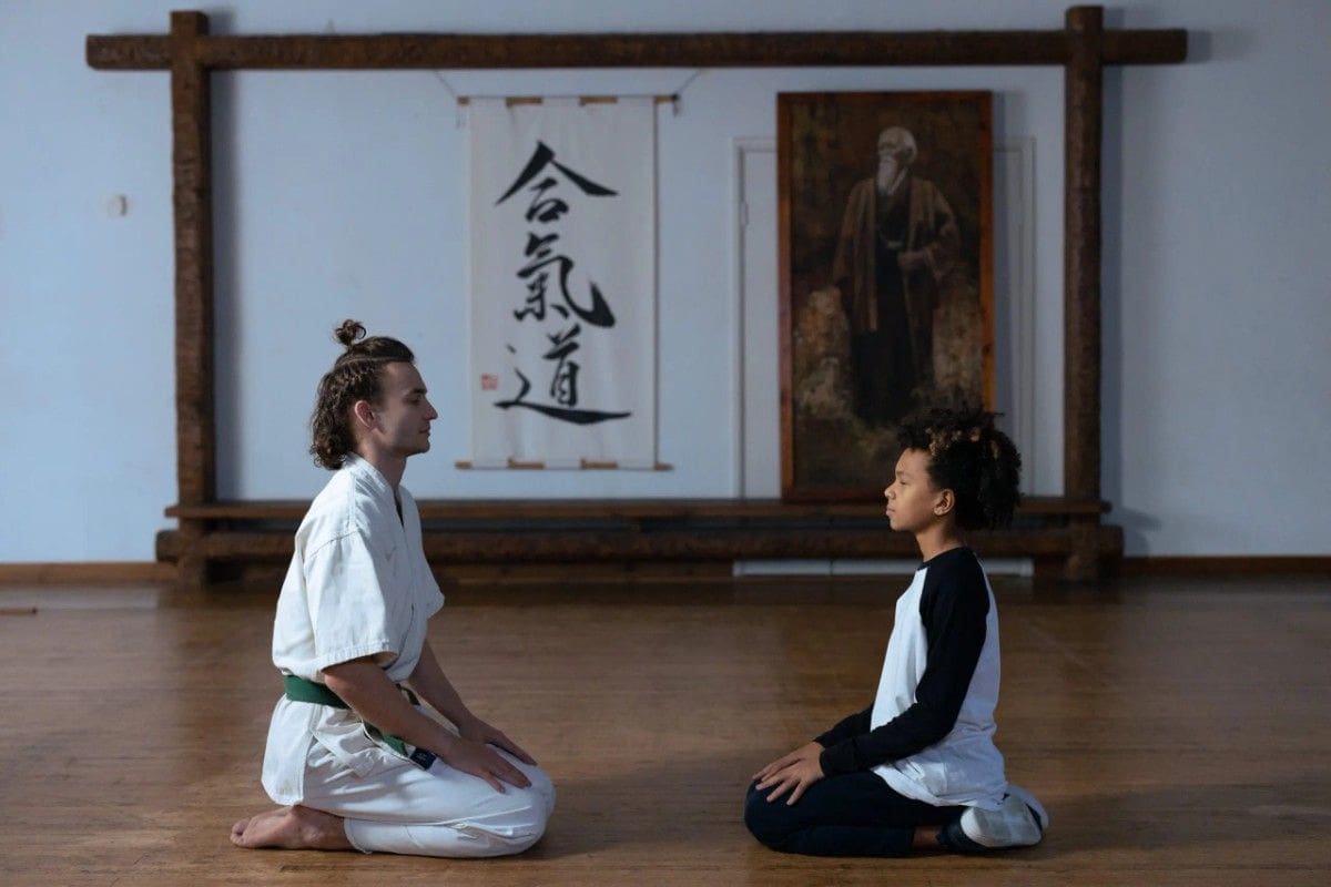 Martial arts instructor and student kneeling in meditation pose inside traditional dojo setting.