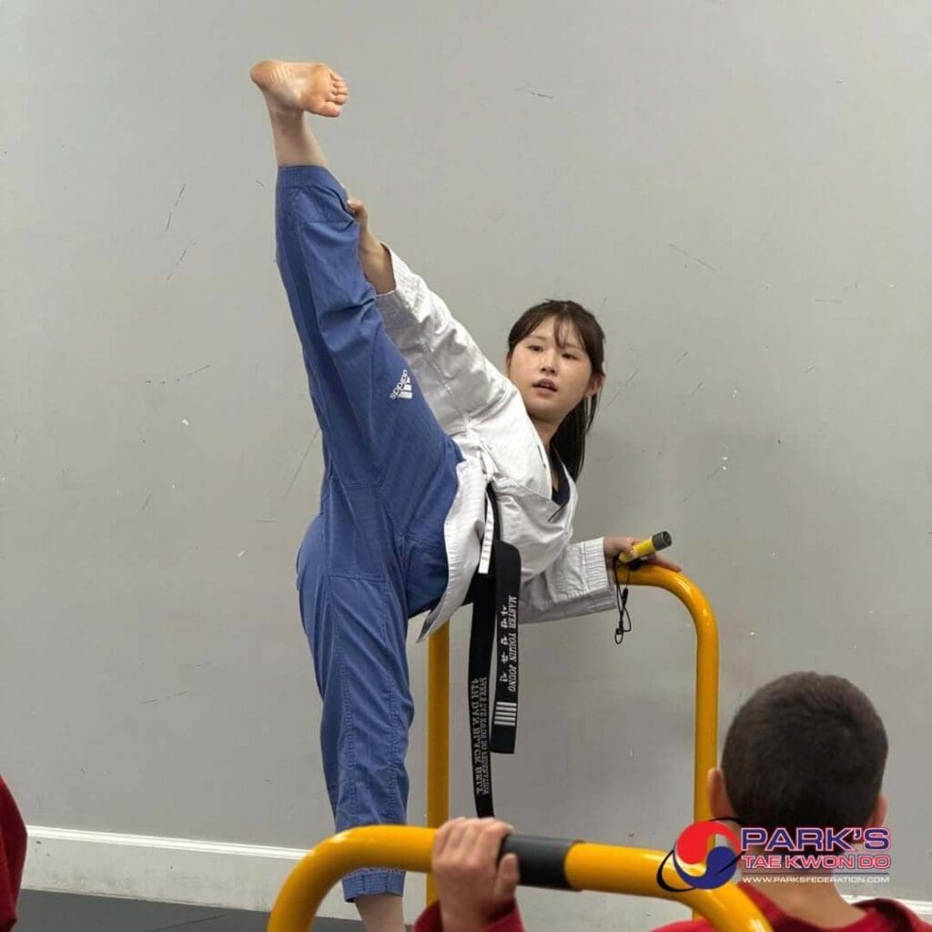Taekwondo student performing a high kick with support during flexibility training in a martial arts class, demonstrating martial arts progression.